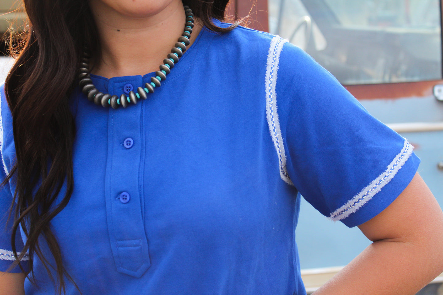 Person wearing a blue shirt with white trim and a necklace, standing outdoors.