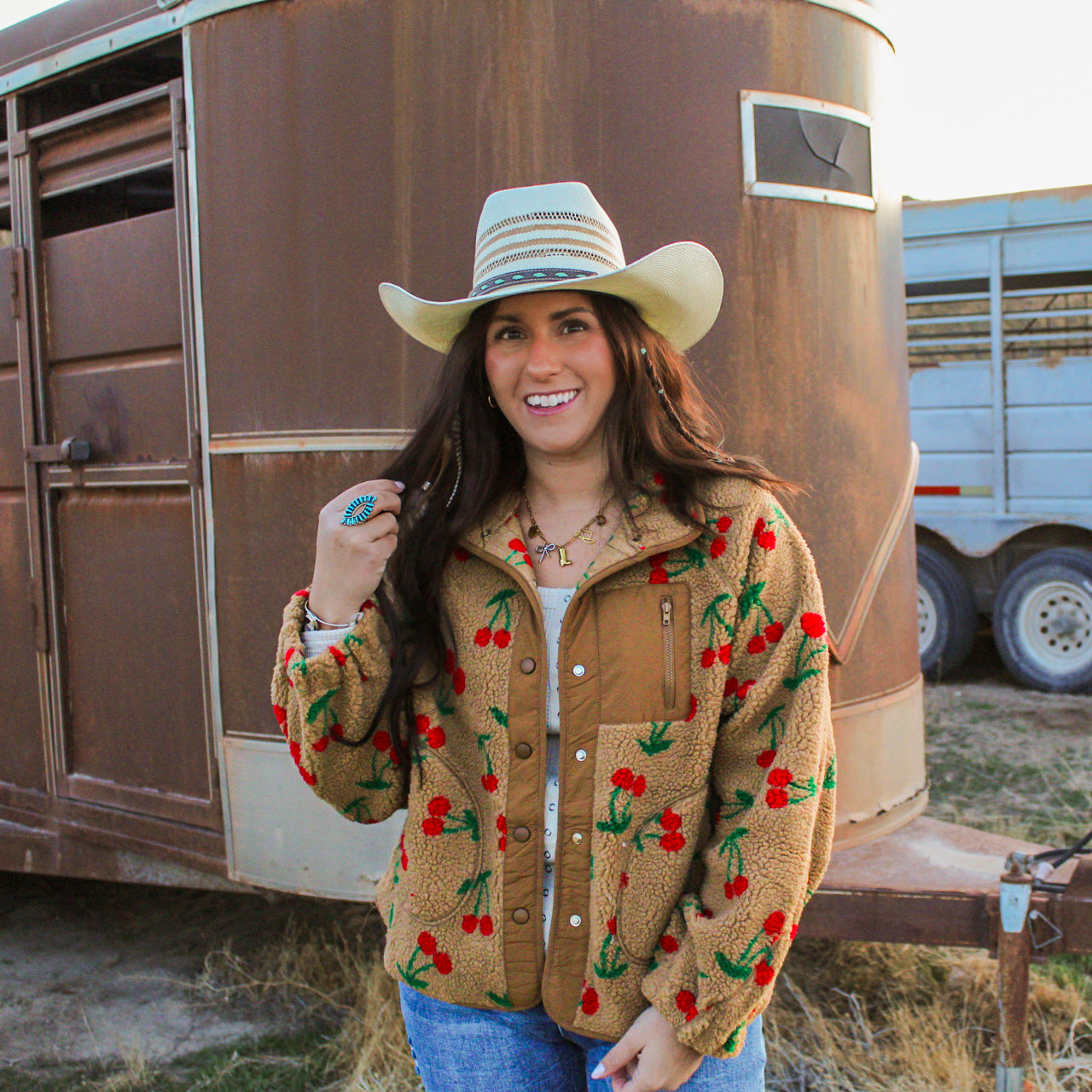 Woman wearing a cowboy hat and a jacket with cherry pattern in front of a trailer.