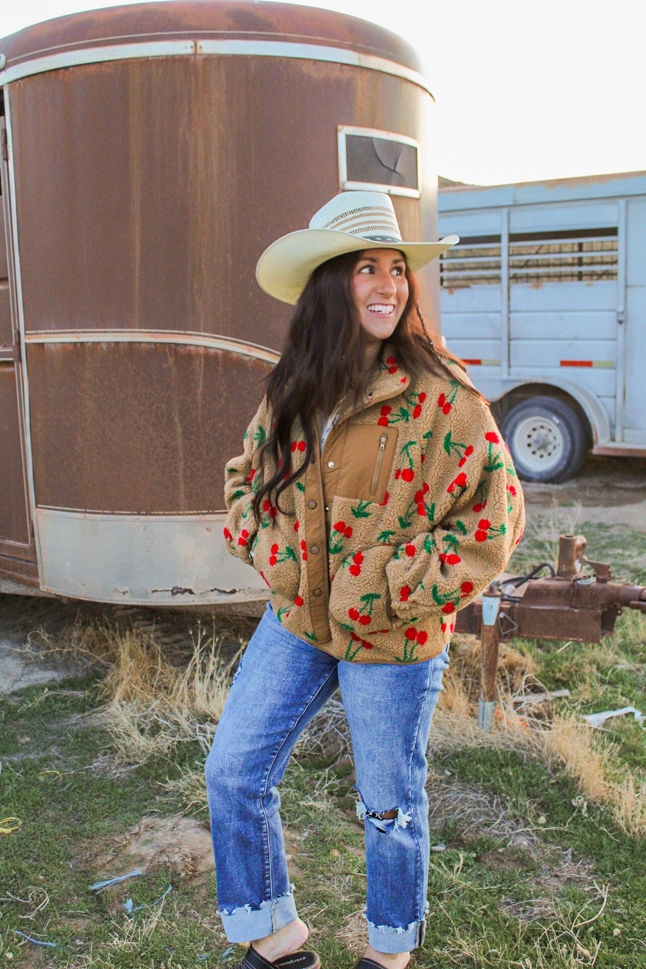 Woman wearing a brown jacket with red patterns, blue jeans, and a white hat standing in front of a trailer.