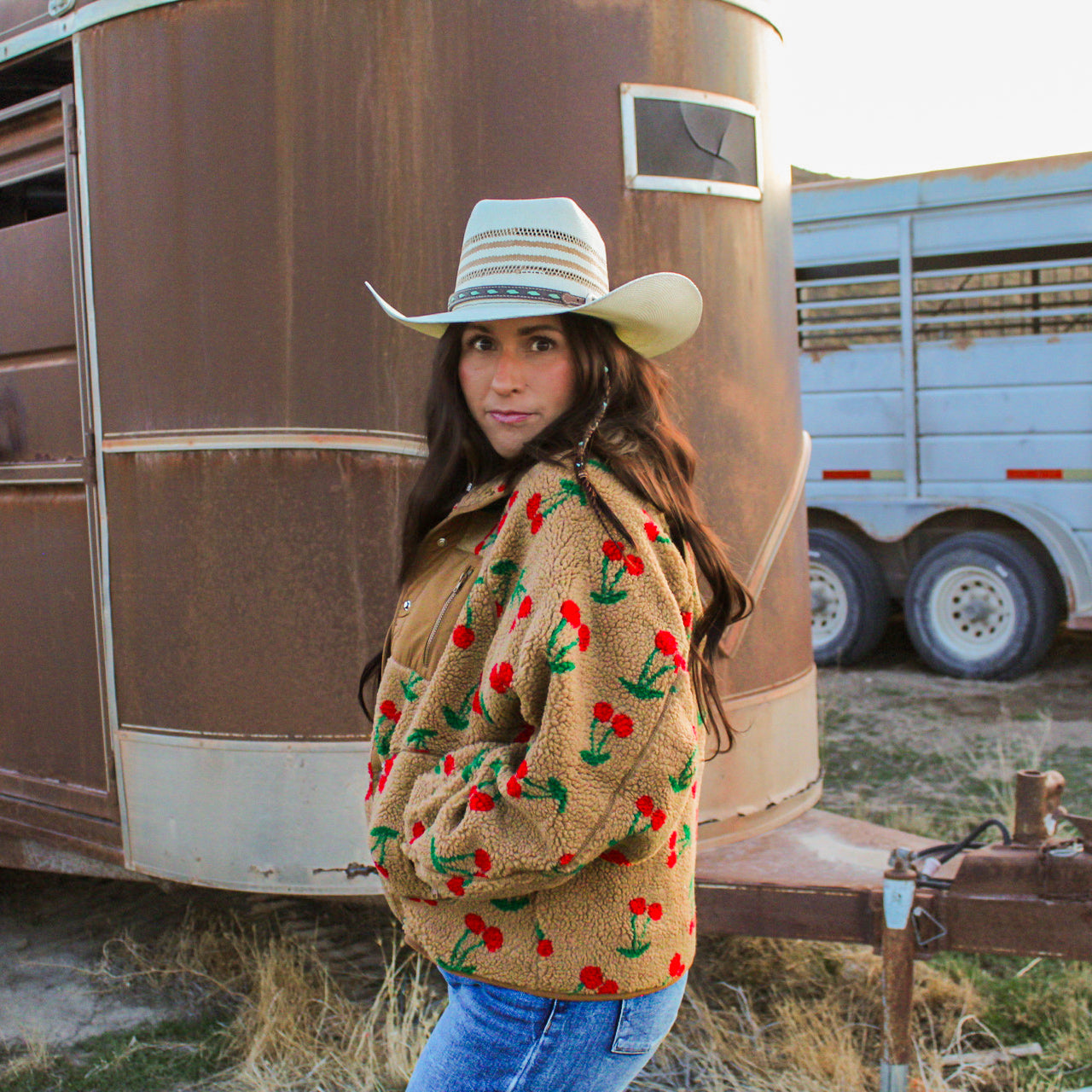 Woman wearing a patterned jacket and cowboy hat standing in front of a rustic trailer.