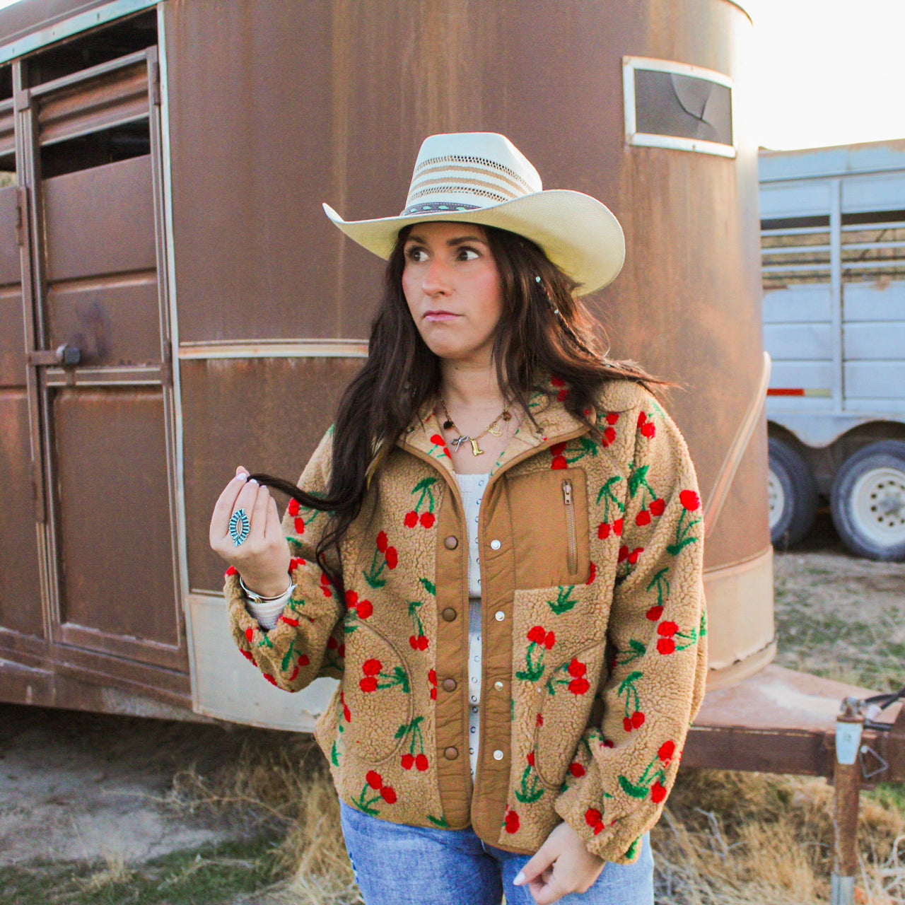 Woman wearing a tan jacket with cherry pattern and cowboy hat in front of a trailer.